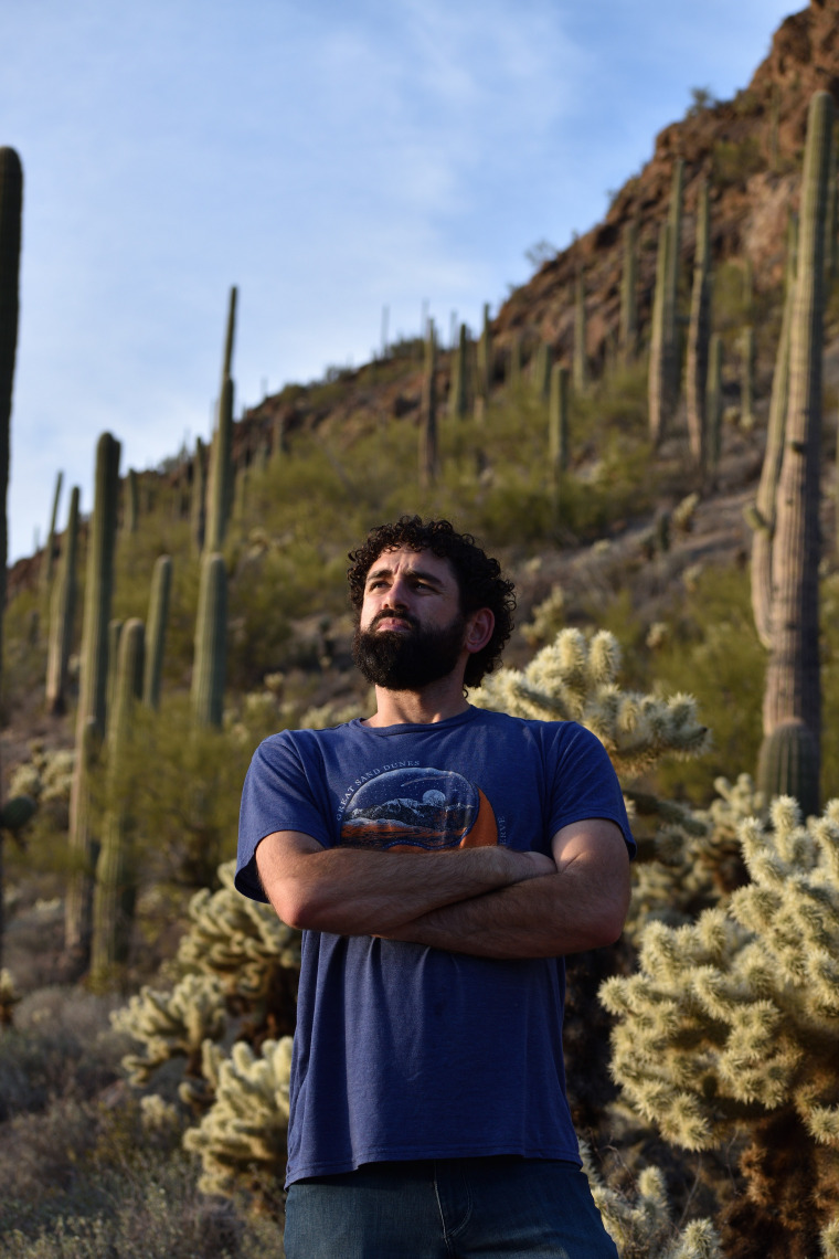 Curly haired man crossing arms looking into the distance in a desert landscape. 